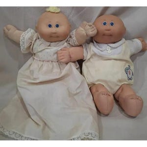 Puede incluir: Dos muñecas Cabbage Patch Kids vintage. Una muñeca lleva un vestido color crema con ribete de encaje, y la otra lleva un pelele amarillo con cuello blanco. Ambas muñecas tienen ojos azules y cuerpos de tela suave.