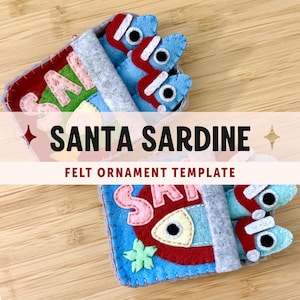 May include: Two felt sardine ornaments featuring blue fish with red Santa hats and white trim. The fish are tucked into grey felt tins with pink lettering that reads SANTA SARDINE. The ornaments include stitched details and small black eyes.