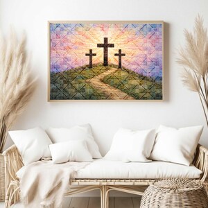 May include: A framed artwork depicts three dark crosses on a hill with a path leading to them. The background features a colorful sunset with shades of pink, yellow, and blue. The artwork is in a light-colored wooden frame.