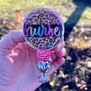 May include: A round badge reel with the word "nurse" in a colorful script. The badge has a leopard print background and is accented with pink and iridescent beads.