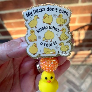 May include: A badge reel featuring a glittery, scalloped-edge acrylic topper with yellow duck illustrations and the text "My Ducks don't even know what a row is". Below the topper is an orange glitter bead and a small yellow rubber duck charm.