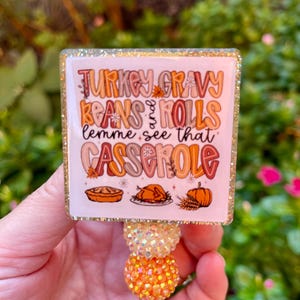 May include: A glittery badge reel with a Thanksgiving theme. The square top features the text "TURKEY GRAVY BEANS & ROLLS lemme see that CASSEROLE" with illustrations of a pie, turkey, and pumpkin. The reel has orange and yellow beaded accents.