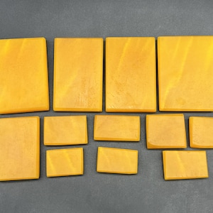May include: A collection of eleven rectangular blocks in varying sizes, all featuring a smooth, solid mustard yellow surface with subtle tonal variations and a matte finish, arranged in two rows on a dark grey surface.