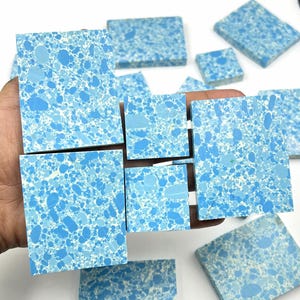 May include: A collection of blue and white speckled square and rectangular tiles. The tiles are held in a hand and are of varying sizes. The speckled pattern is a mix of light and dark blue, creating a unique design.