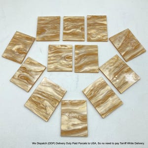 Puede incluir: Once piezas rectangulares con acabado brillante, que presentan un patrón de remolinos en tonos crema y marrón dorado. Cada pieza tiene una superficie lisa y plana con esquinas redondeadas, dispuestas en una formación simétrica.