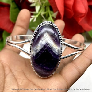 Peut inclure: Un bracelet manchette en argent orné d'une grande pierre d'améthyste ovale présentant des bandes en chevron violettes et blanches. La pierre est sertie dans un bord en argent torsadé, et le bracelet se compose de deux bras en métal argenté.