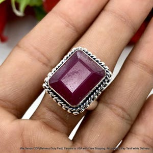 May include: A rectangular, deep red gemstone ring set in a polished silver band. The stone features a faceted surface and is surrounded by a decorative twisted rope-style metal border. The silver band has a smooth, rounded finish on the sides.