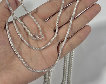 Puede incluir: Un collar de cadena plateada con un diseño tejido texturizado. El collar se sostiene en una mano, con el cierre visible. El texto "DESIGN 1" está en la esquina superior izquierda. La cadena mide unos 50 cm.