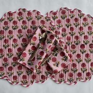 Op de afbeelding: Twee ronde, doorgestikte placemats met geschulpte randen en een roze achtergrond met een herhalend bloemmotief in donkerroze en groen. Een bijpassend gevouwen servet ligt erbovenop, met een zilverkleurige metalen lepel en een zilverkleurige vork.
