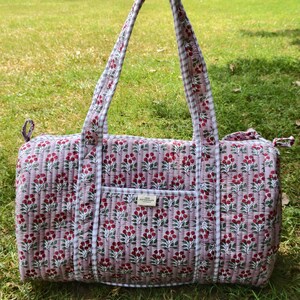 May include: A quilted duffel bag featuring a light pink fabric with a repeating pattern of small red cherries and green leaves. The bag has two long handles and trim in a white and grey gingham check pattern, with a small rectangular label.