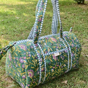 Könnte beinhalten: Eine gesteppte Reisetasche mit grünem Grund und einem Blumenmuster in Rosa, Gelb und Weiß. Die Tasche verfügt über zwei Tragegriffe und eine weiß-grün gestreifte Verzierung sowie ein kleines rechteckiges Etikett auf der Vorderseite.