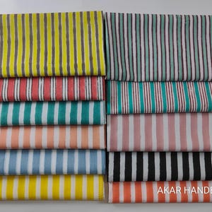 Puede incluir: Conjunto de telas plegadas con rayas verticales en varios colores: amarillo, rojo, turquesa, melocotón, azul, rosa, negro y naranja. El texto "AKAR HANDBLOCK" es visible. Las telas están apiladas ordenadamente.