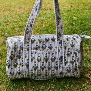 May include: A quilted duffel bag featuring a light grey background with a repeating floral pattern in shades of teal and mustard yellow. The bag includes two long shoulder straps, a zippered top closure, and a small exterior pocket on the front.