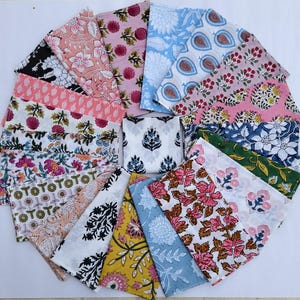 May include: A collection of square fabric napkins arranged in a circular fan pattern, featuring a variety of block-printed floral and botanical designs in colours including pink, blue, yellow, green, black, and white on a white background.