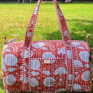 May include: A quilted duffel bag featuring a vibrant orange and white floral block print pattern. The bag includes two long handles with a red and white striped design, a top zipper closure, and a small rectangular label on the front pocket.