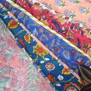 May include: A close-up of several colorful quilted fabric pieces with floral patterns. The fabrics feature a variety of colors, including red, blue, pink, and green, with intricate designs. The quilting adds texture and dimension to the textiles.