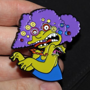 May include: Purple and yellow cartoon character with multiple eyes and a blue dress. The character has a large purple afro with white and blue circles. The character is a pin.