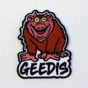 Peut inclure: Patch brodé représentant un personnage de dessin animé avec une fourrure rouge, des yeux verts et un large sourire. Le personnage est assis sur un nuage blanc avec le mot "GEEDIS" écrit en noir.