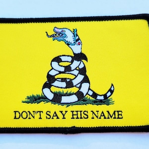 Op de afbeelding: Een gele patch met een zwart-witte slang met een menselijk hoofd. De slang is opgerold en heeft een kroon op zijn kop. De tekst "DON'T SAY HIS NAME" staat onder de slang.