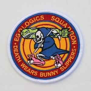 Puede incluir: Un parche redondo con borde blanco y un diseño rojo, amarillo y azul. En el centro, un esqueleto con una túnica azul que lleva un torpedo verde y amarillo. El texto alrededor del borde dice "Death Wears Bunny Slippers - Ecologics Squadron".