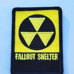 Può includere: Toppa ricamata gialla e nera con un simbolo di radiazione stilizzato e il testo "Fallout Shelter".