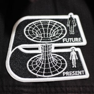 May include: Black and white embroidered patch featuring a stylised image of a black hole. The patch has the words "FUTURE" and "PRESENT" on it.