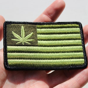 May include: Green and black embroidered patch with a marijuana leaf and an American flag design.
