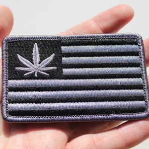 May include: A black and silver embroidered patch featuring a marijuana leaf and an American flag design.