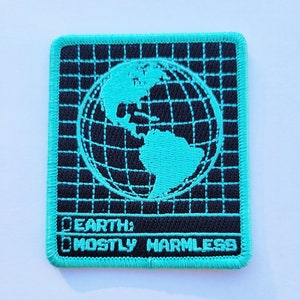 Könnte beinhalten: Ein gestickter Patch in Türkisblau mit einem schwarzen Rastermuster und einem türkisblauen Globus in der Mitte. Der Text "EARTH: MOSTLY HARMLESS" steht unter dem Globus.