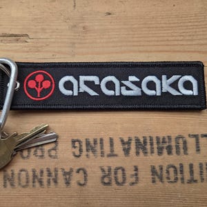 Peut inclure: Porte-clés rectangulaire noir avec le mot ARASAKA en blanc et un logo rouge et blanc. Le porte-clés est attaché à un mousqueton argenté avec plusieurs clés. Le fond est une surface en bois.