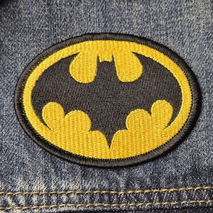 May include: An embroidered patch featuring the iconic Batman logo in yellow and black. The oval-shaped emblem displays a stylized bat silhouette centered within a vibrant yellow border, all stitched onto a textured blue denim fabric background.