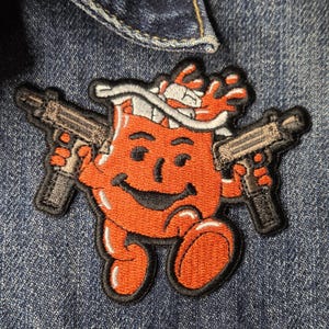 May include: An embroidered patch featuring a round, orange, smiling character with a white headband, holding a black submachine gun in each hand. The patch has a thick black border and is attached to a blue denim fabric background.