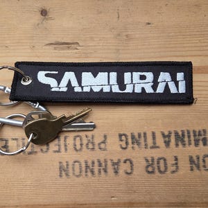 以下が含まれることがあります： 白い「SAMURAI」の文字が刺繍された黒い長方形のキーホルダー。タグはシルバーのカラビナと2つのキーに付いています。背景は木製の表面です。