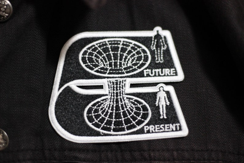 May include: Black and white embroidered patch with a stylized image of a black hole. The patch has the words "FUTURE" and "PRESENT" on it.