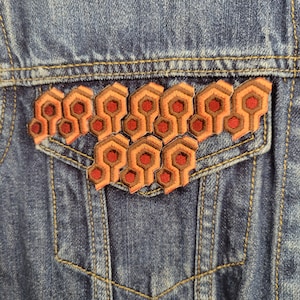 Może przedstawiać: Naszywkę haftowaną w kolorze czerwonym i pomarańczowym z ikonicznym wzorem dywanu z hotelu "Overlook" z filmu "Lśnienie". Naszywka jest przyszyta do niebieskiej dżinsowej kurtki.