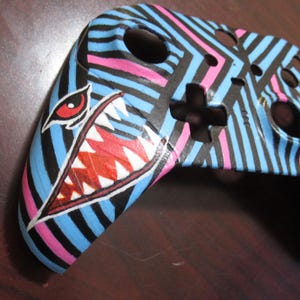 Puede incluir: Una piel para mando de videojuegos a rayas azules y rosas con un diseño de dientes de tiburón rojos y blancos.