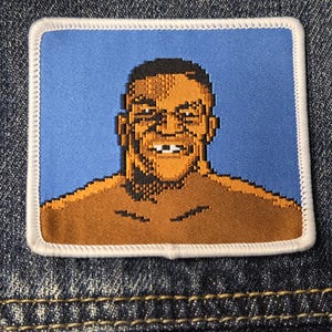 May include: A rectangular embroidered patch featuring a pixelated graphic of a person with a dark complexion, short dark hair, and a visible gap in their front teeth. The figure is set against a light blue background with a white border.