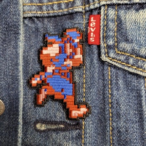 May include: A blue and red pixelated character embroidered on a denim jacket with a Levi's patch.