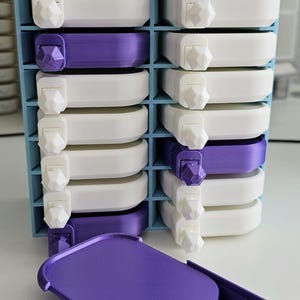 May include: A light blue organizer with multiple compartments holding white and purple containers. Each container has a geometric-shaped handle. One purple container is open, revealing its interior.