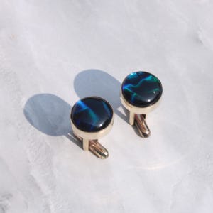 May include: A pair of gold-toned cufflinks with round, iridescent, dark blue and green centers. The cufflinks are set against a white background, with a shadow cast to the left.