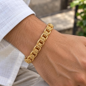 Puede incluir: Una pulsera de tono dorado con un diseño de cadena bizantina compleja y multicapa. Los eslabones metálicos están interconectados en un patrón repetitivo e intrincado que crea una banda gruesa y texturizada alrededor de la muñeca.