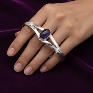 Può includere: Un braccialetto argentato con una gemma ovale, viola scuro al centro. Il braccialetto ha un design diviso con accenti decorativi su ciascuna estremità. Il gioiello è esposto su uno sfondo viola scuro.