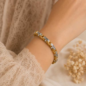 May include: A two-tone bracelet featuring a repeating pattern of interlocking gold-colored links and silver-colored rectangular segments. The design mimics the appearance of a bicycle chain, creating a structured and metallic aesthetic on the wrist.