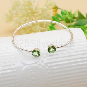 May include: A silver-toned bangle bracelet with two round, light green gemstones. The bracelet has a simple, open design. The gemstones are set at the ends of the bracelet. The background is white with some green foliage.