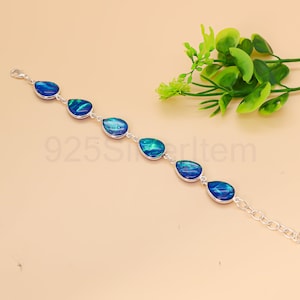 Puede incluir: Una pulsera de plata con ópalos azules y verdes en forma de lágrima. Los ópalos están engarzados en una cadena de plata, con un cierre de mosquetón. La pulsera se exhibe sobre un fondo neutro con follaje verde.