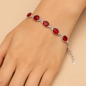Puede incluir: Una pulsera plateada con piedras preciosas rojas brillantes de forma ovalada. La pulsera está compuesta por una serie de piedras enlazadas, con una delicada cadena y un cierre ajustable. Las piedras están engastadas en metal plateado.