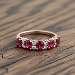 Peut inclure: Bague en or rose ornée d'une série de pierres précieuses rouges vives de forme ovale, alternant avec de plus petites pierres précieuses claires et rondes. La bague est posée sur une surface en bois rustique.
