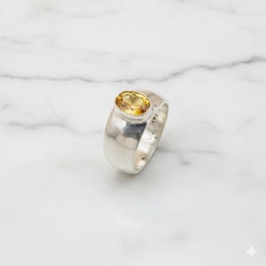 May include: A silver ring featuring a large, oval yellow gemstone. The ring has a wide band and a polished finish, set against a white marble surface. The gemstone is a vibrant yellow colour.
