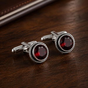 May include: A pair of round cufflinks featuring a faceted deep red gemstone set in a polished silver-tone metal frame. The circular setting is bordered by a decorative twisted rope texture, and each cufflink includes a standard hinged toggle back.