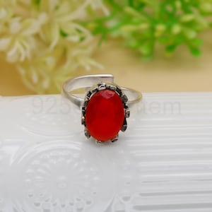 May include: A silver ring featuring a large, oval-shaped, faceted red gemstone. The gemstone is set in a decorative, textured bezel. The ring has a simple, open band design.